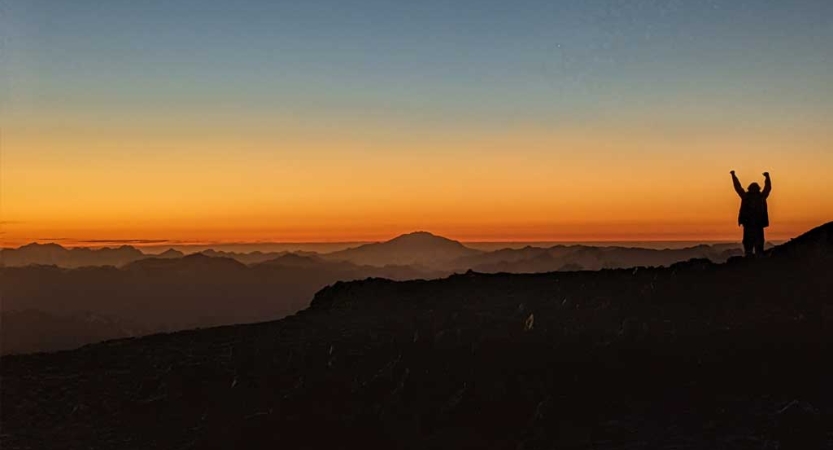 A person raises their hands in celebration on an overlook above a vast mountain range. The sky appears in shades of orange and yellow as the sun either sets or rises. 